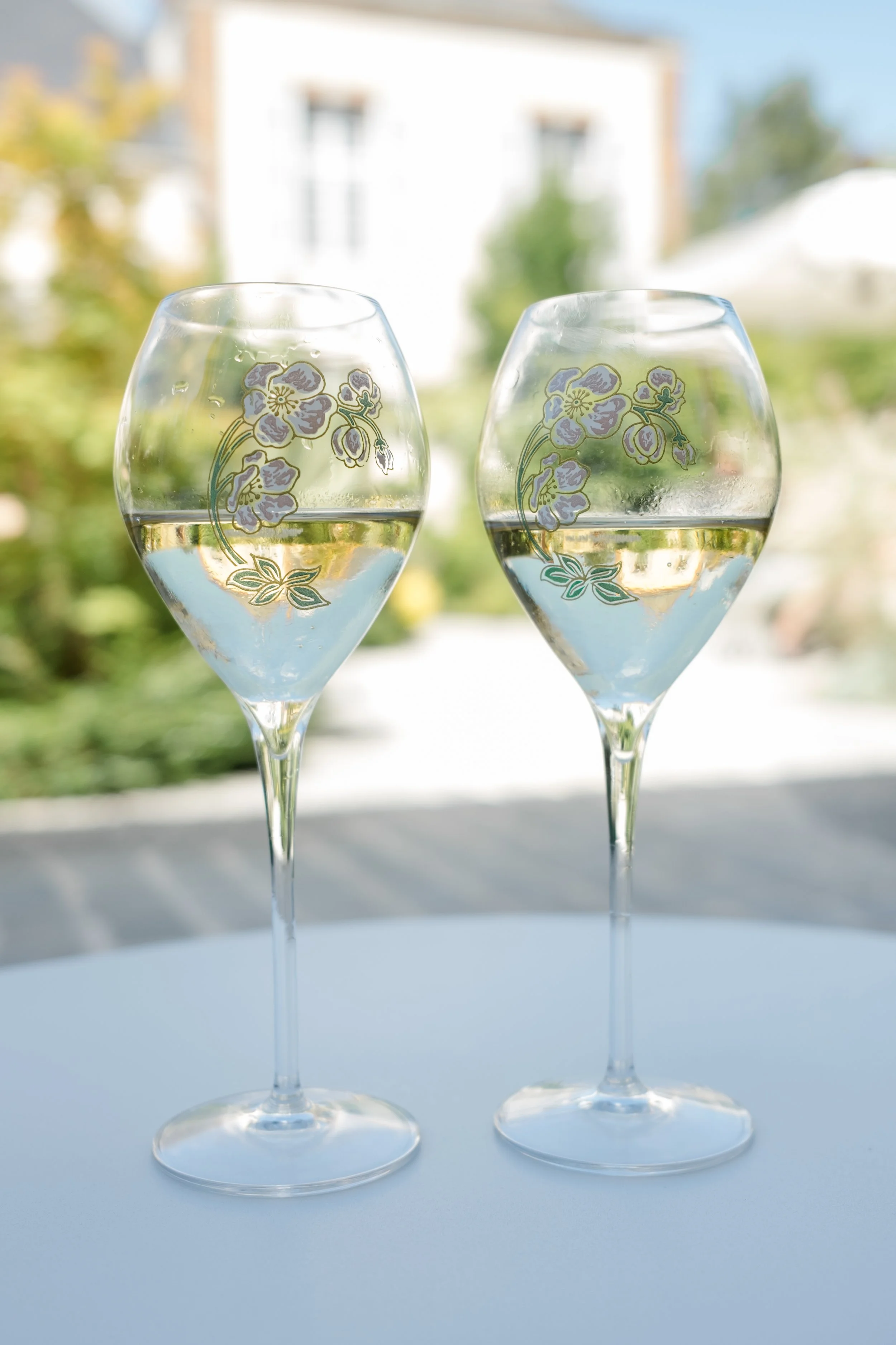 Two wine glasses with floral designs on a white surface, outside with a blurred background of trees and buildings.