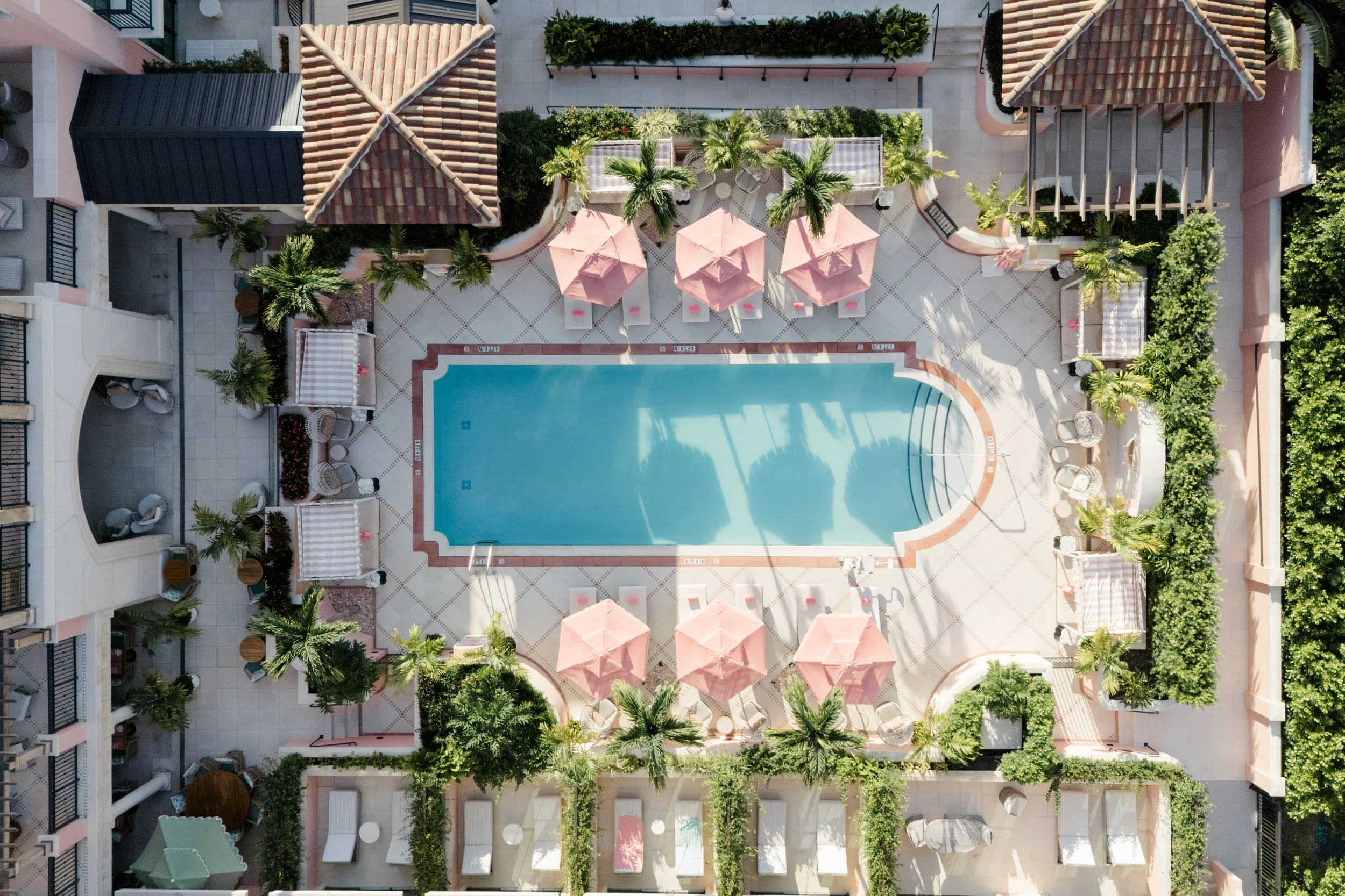 An aerial view of a pool area surrounded by pink umbrellas, white lounge chairs, green palm trees, and surrounding buildings with balconies.