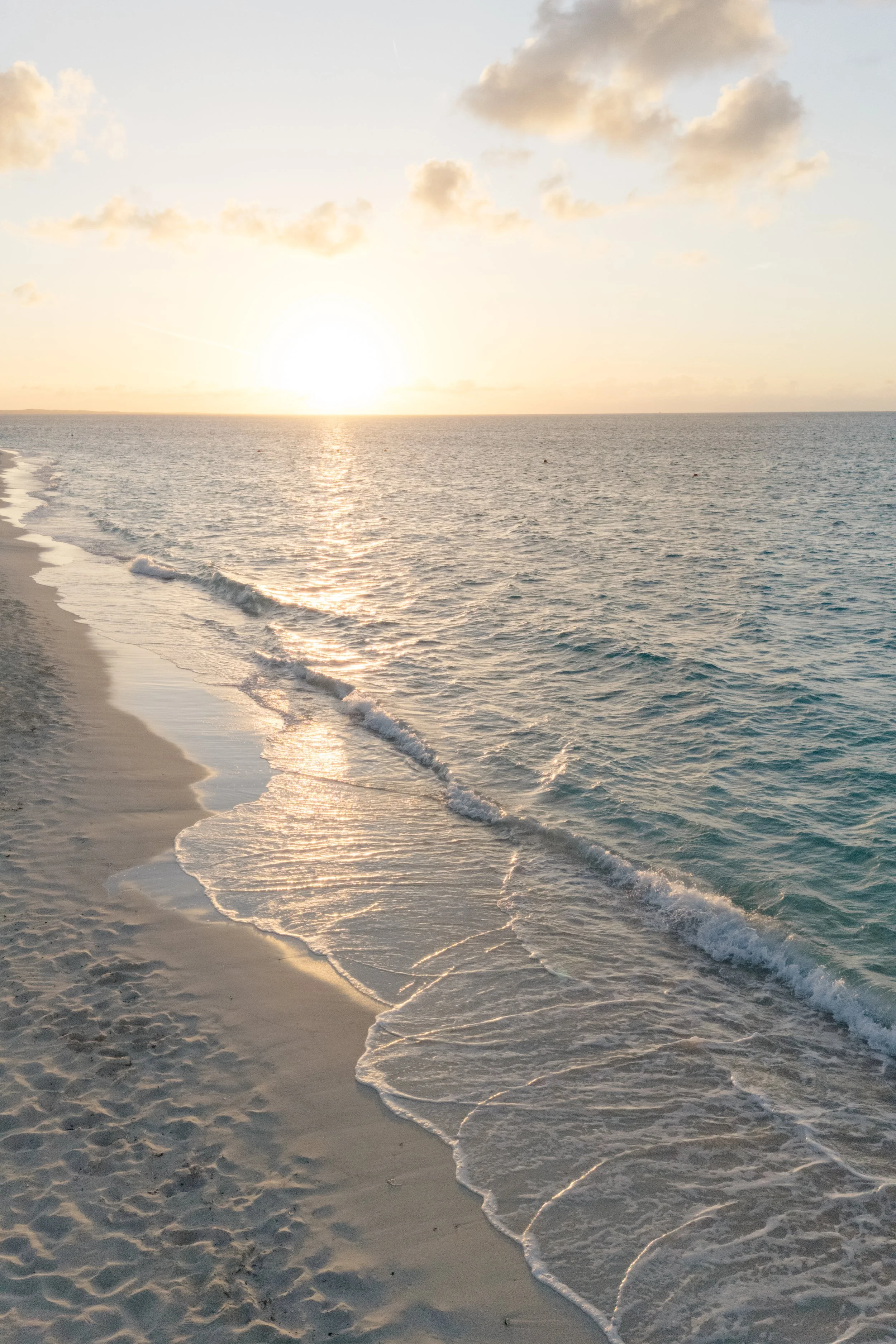 A sandy beach during sunset with gentle waves lapping on the shore and a few clouds in the sky.