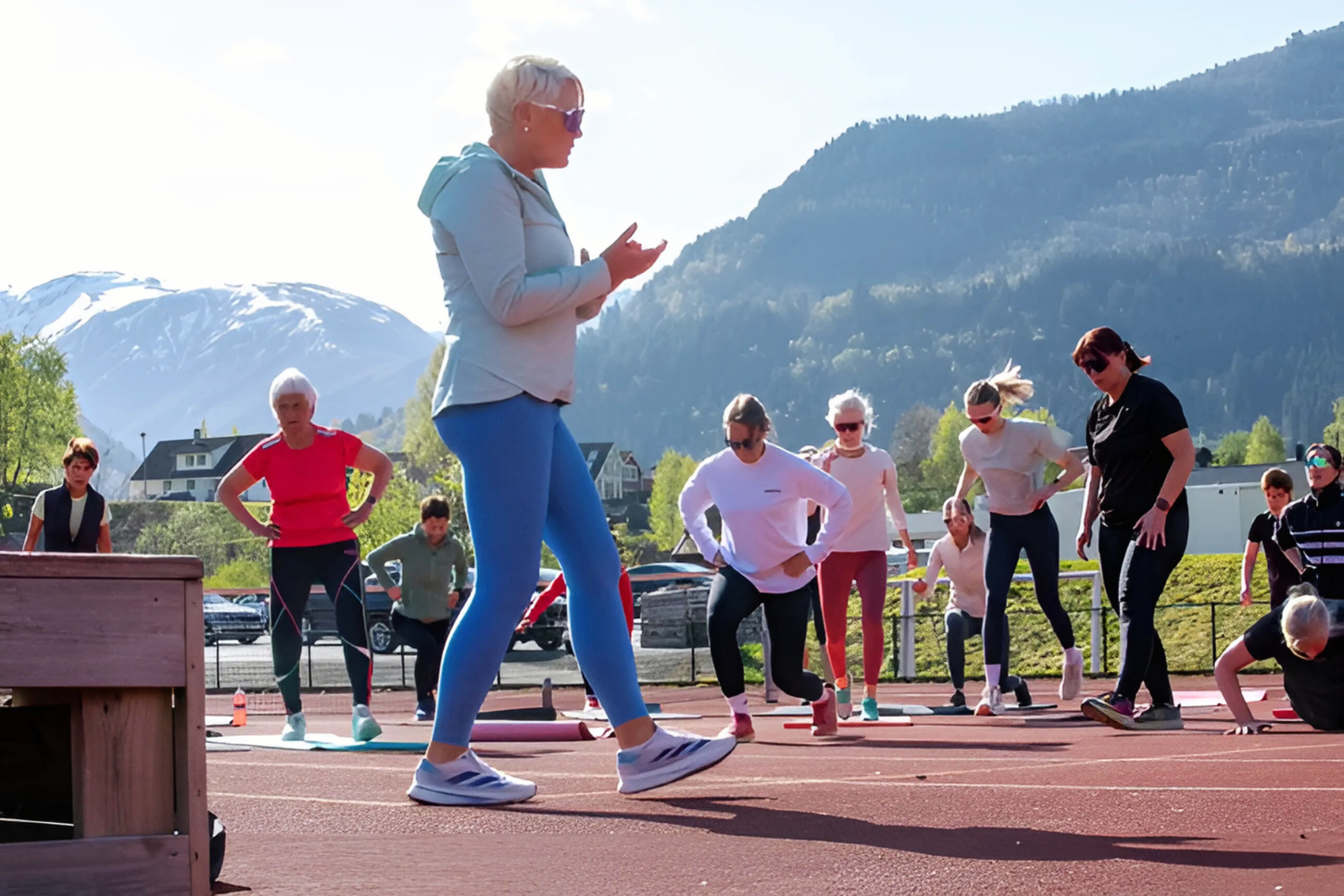 En gruppe mennesker deltar i en utendørs treningsøkt med fjelllandskap i bakgrunnen. En person gir instruksjoner mens de andre følger med og gjør øvelser på en løpebane.