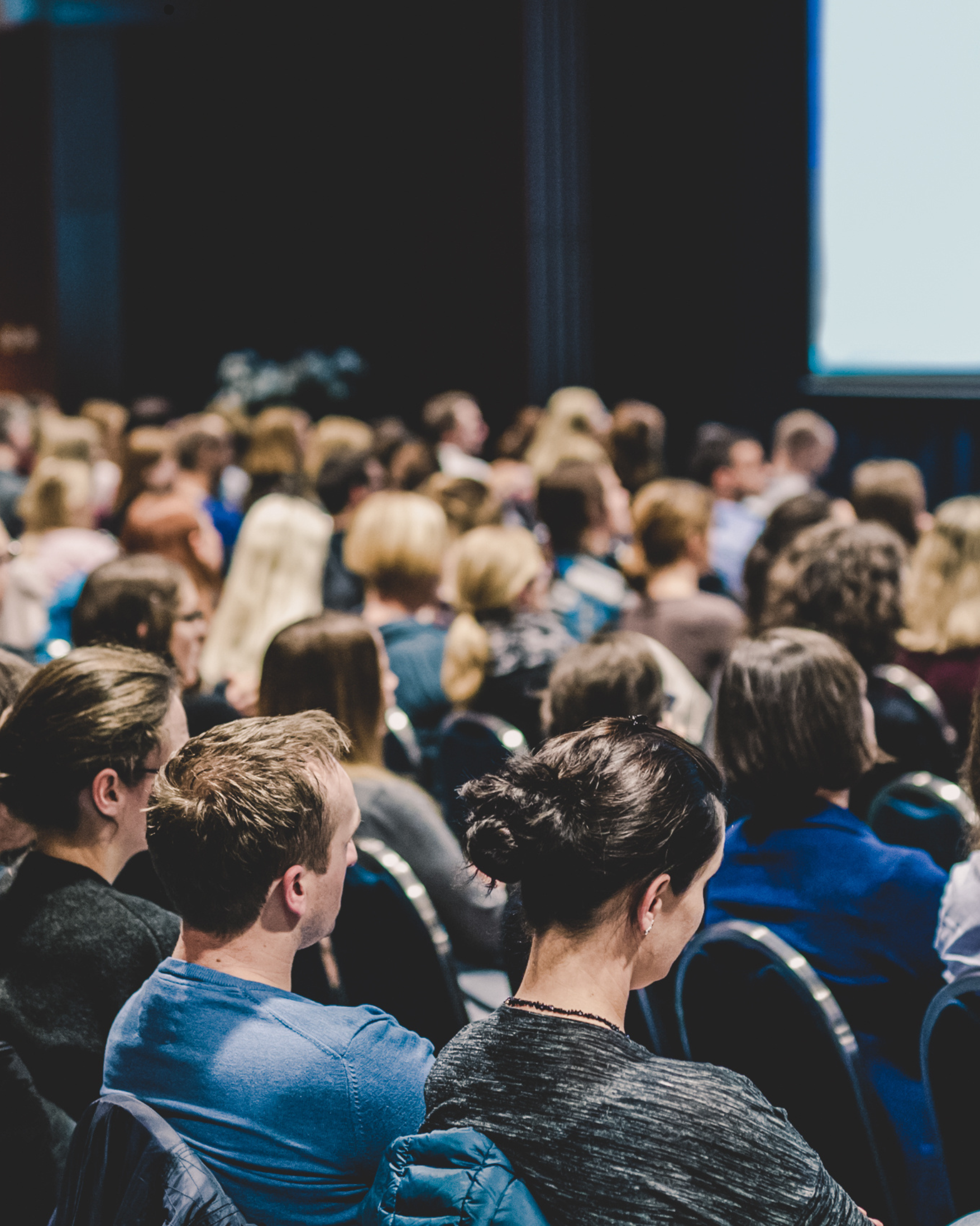 Folk sitter på en konferanse eller forelesning, sett bakfra, med fokus på publikum og en projisert skjerm i bakgrunnen.