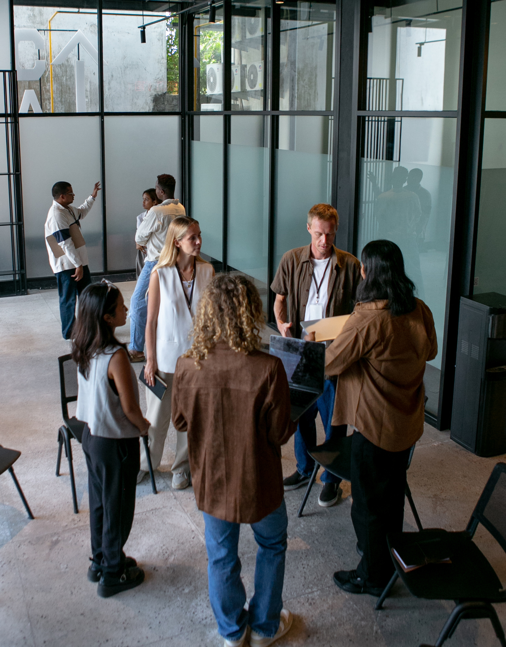 En gruppe mennesker står i et moderne kontorlokale med store glassvegger og stoler. Flere personer er opptatt med forskjellige aktiviteter, som å snakke med hverandre og bruke en bærbar datamaskin. Noen står ved vinduene og ser ut. Det er en uformell og samarbeidsorientert atmosfære.