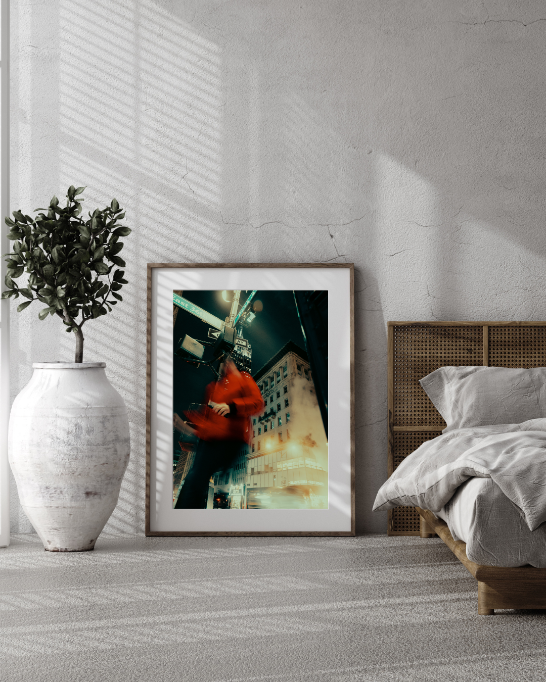 A framed cityscape photo with a blurred person in a red coat standing on a street corner, leaning against a building at night, next to a large potted plant on a light-colored carpeted floor beside a bed with white bedding.