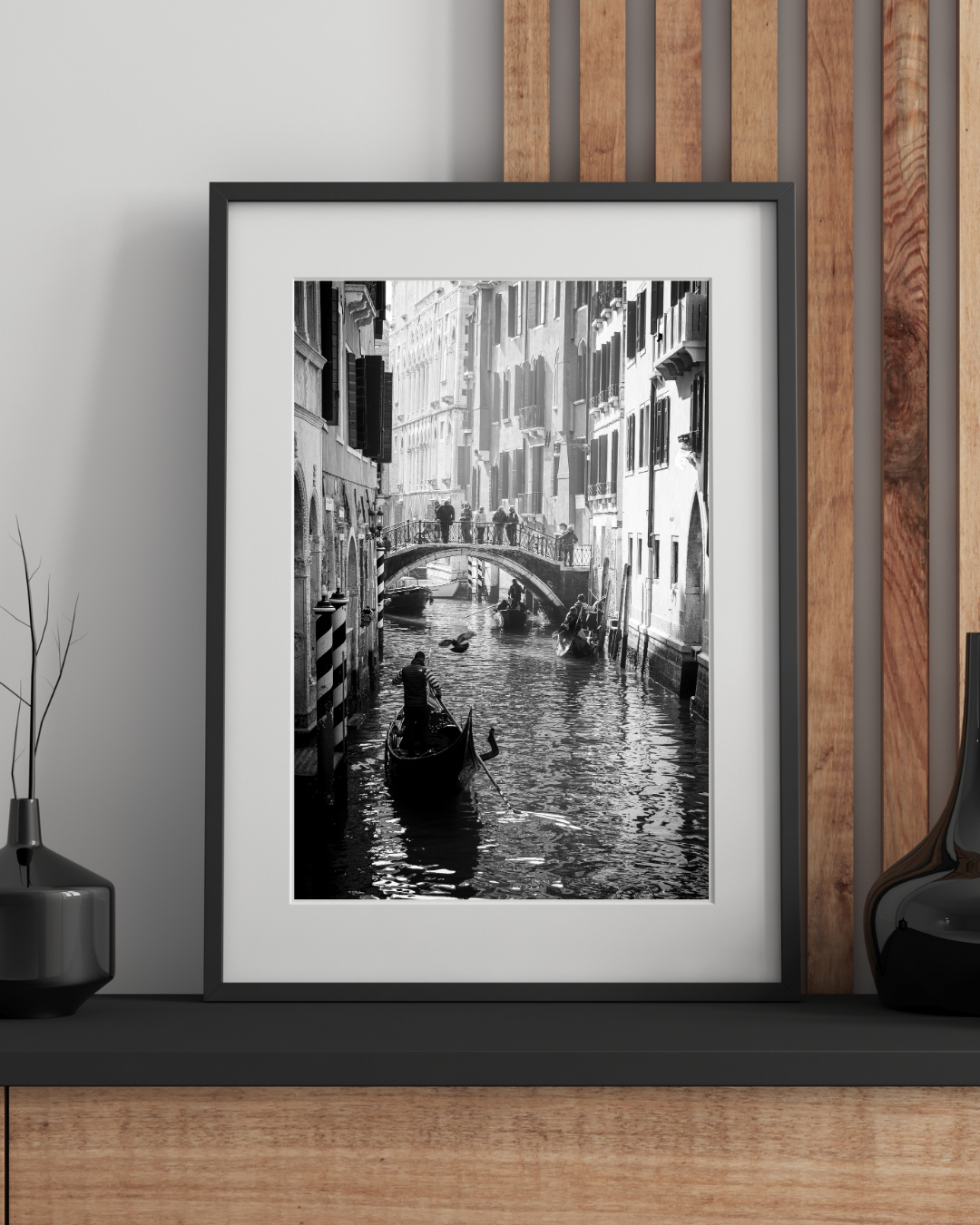 Black-and-white photograph of a canal in Venice, Italy, with gondolas and historic buildings, framed and displayed on a shelf.
