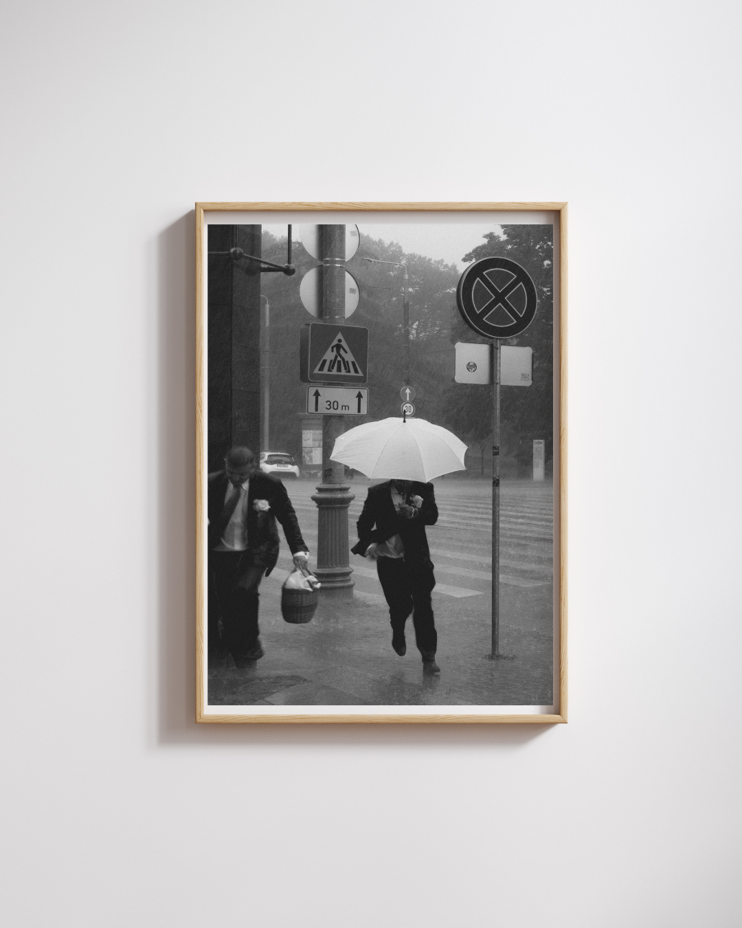 Black and white photograph of two people walking in the rain with an umbrella, crossing a city street near traffic signs and a lamppost.