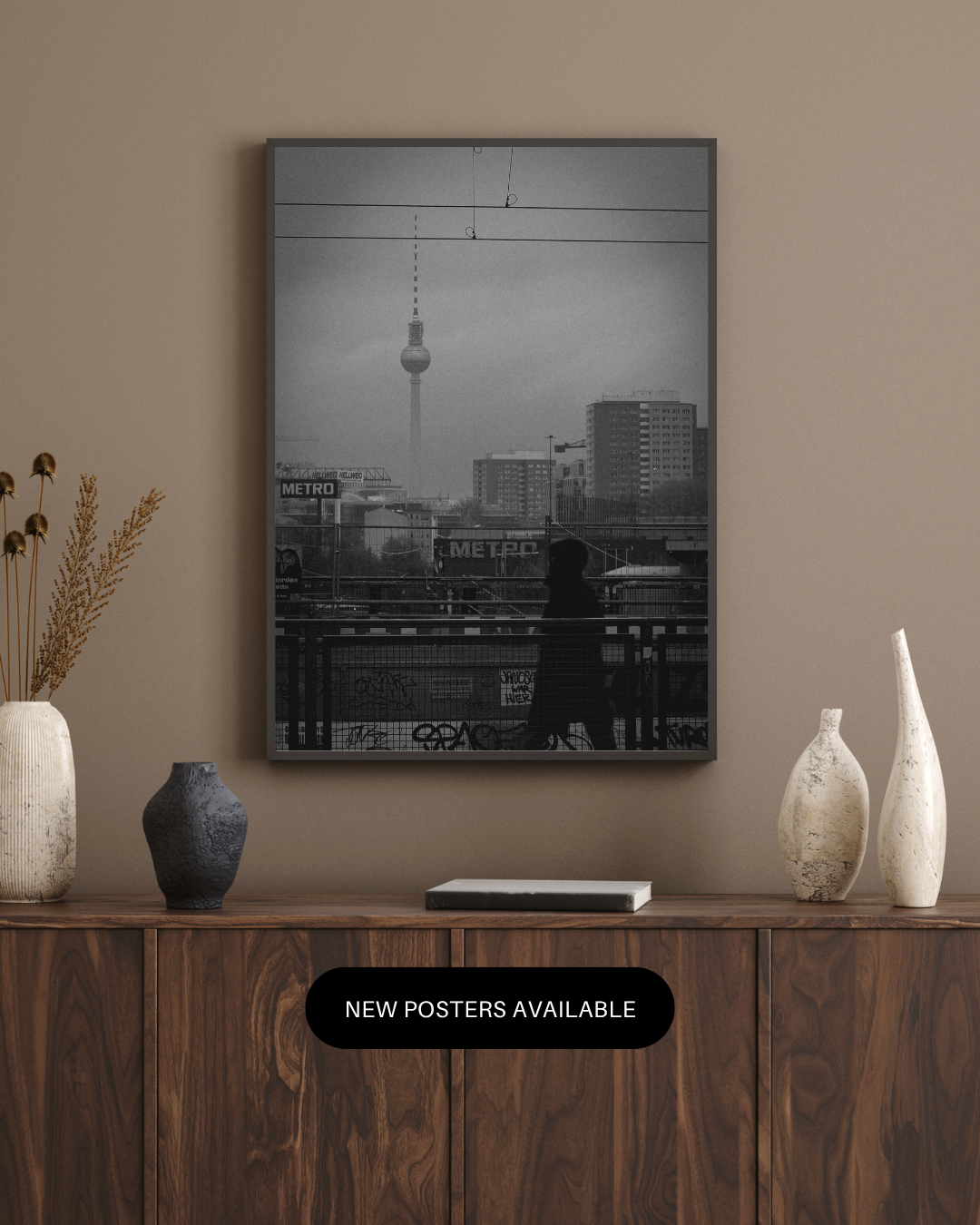 Black and white cityscape photo of Berlin, Germany, with the Berlin TV Tower in the background. A person is sitting on a bench on a bridge, facing away, with graffiti on the railing and metro signs visible.