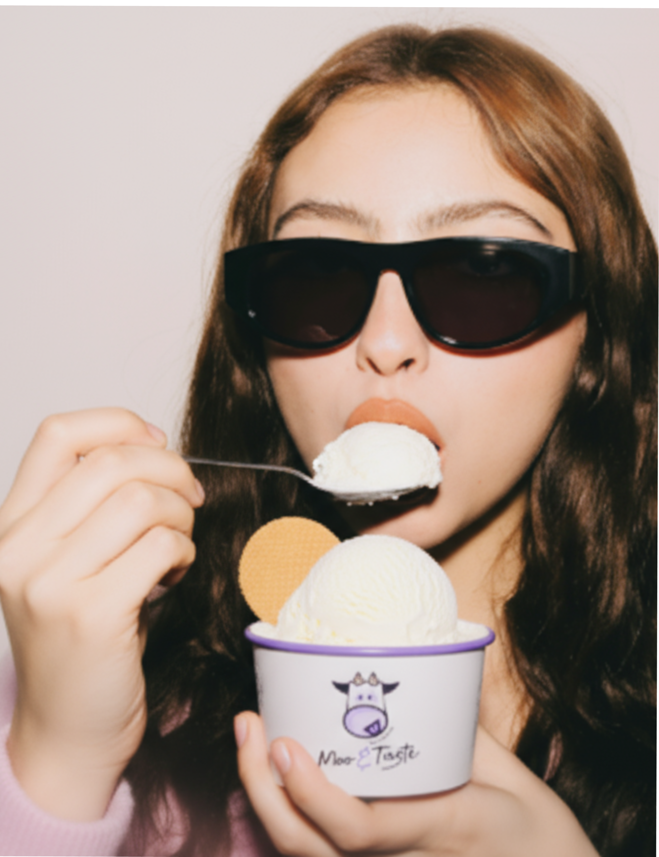 Woman with long brown hair wearing black sunglasses eating ice cream from a white cup with a cow illustration and the words 'Moo & Taste' on it.