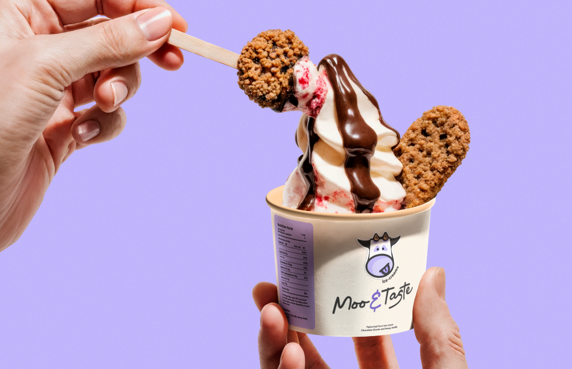 A person holds a wooden stick with a round, breaded ice cream treat above a cup of soft serve ice cream. The cup has a cow logo and the text 'Moo E Taste' on it. The ice cream is topped with chocolate syrup and contains two cookie-like treats.