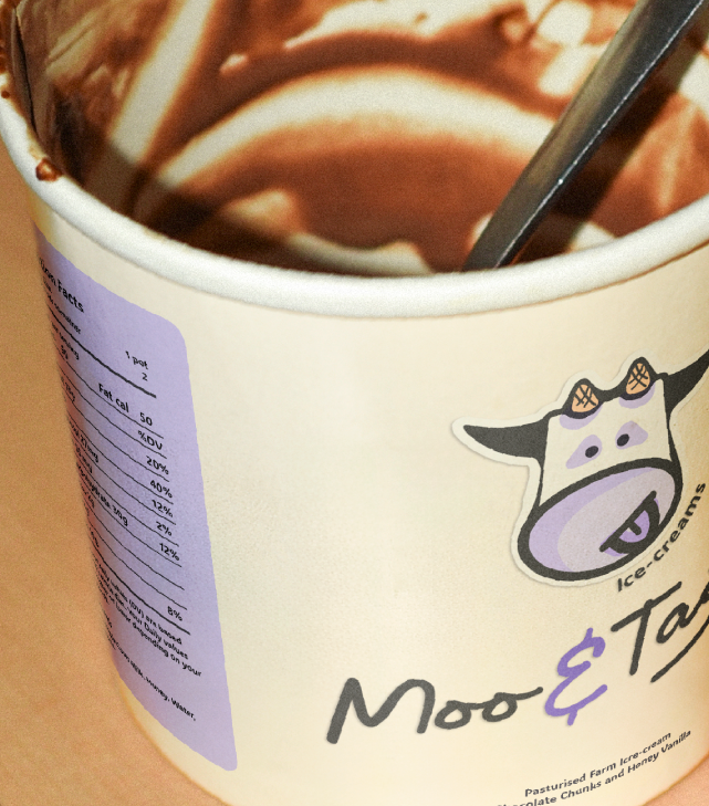 A cup of chocolate ice cream with brown and white swirls, with a spoon inside. The cup features a cow cartoon with ice cream on its head.