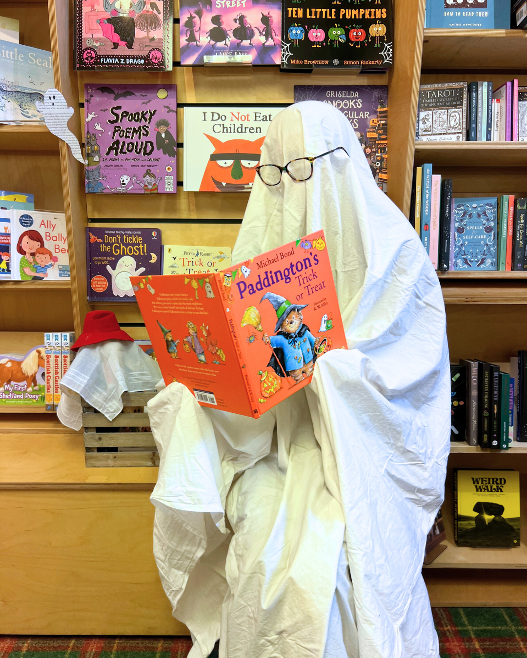 A Halloween-themed display in a bookstore features a figure made of white cloth draped over a person, wearing glasses, holding a copy of 'Paddington's Trick or Treat' with a background of children’s books on shelves.