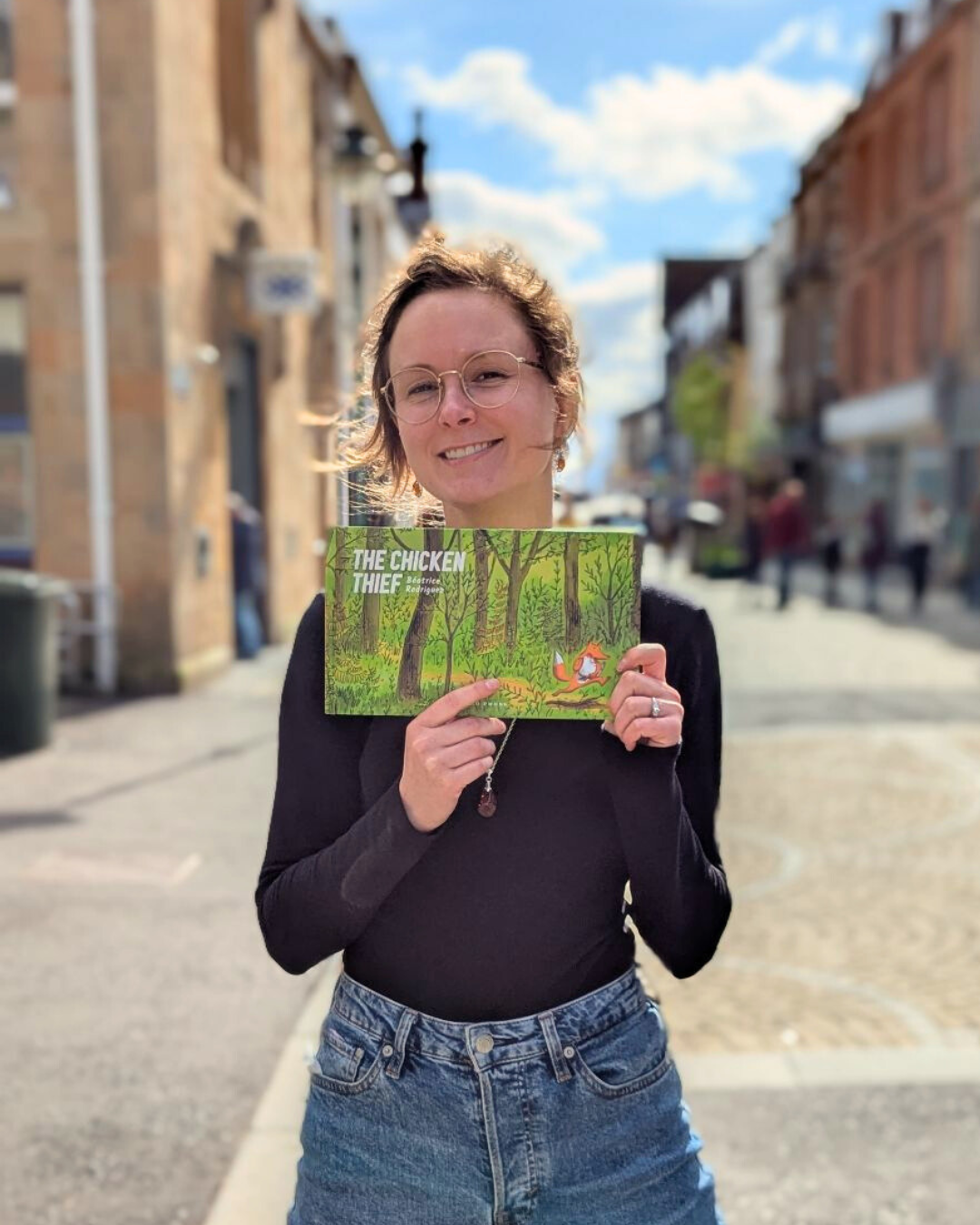 Woman with short hair and glasses smiling outdoors on a city street, holding a children's book titled "The Chicken Thief" by Beatrice Rodriguez.