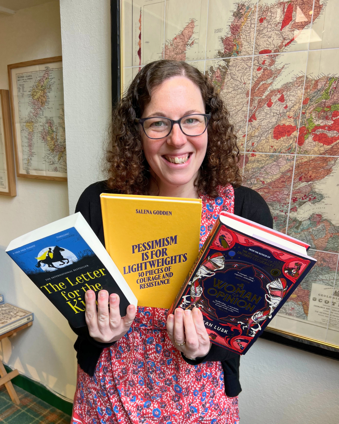 A smiling woman with curly brown hair and glasses holding four books in front of a framed map. The books are titled 'The Letter for the King,' 'Pessimism is for Lightweights,' 'A Woman's Opinion,' and one partially visible.