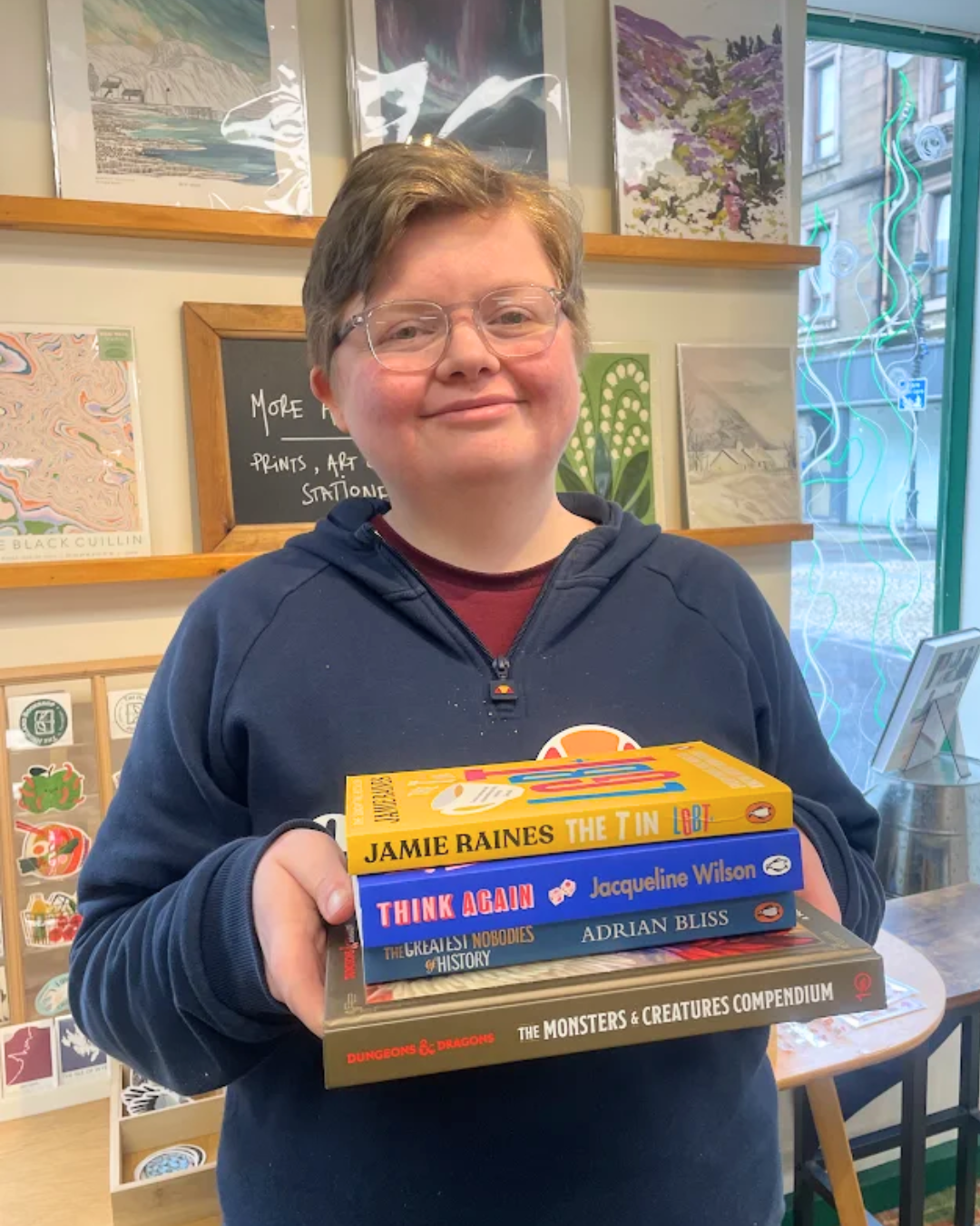A young girl with glasses and long wavy blonde hair smiling outdoors, holding up a book titled 'The Monsters & Creatures Compendium' from Dungeons & Dragons, in front of green leafy plants.