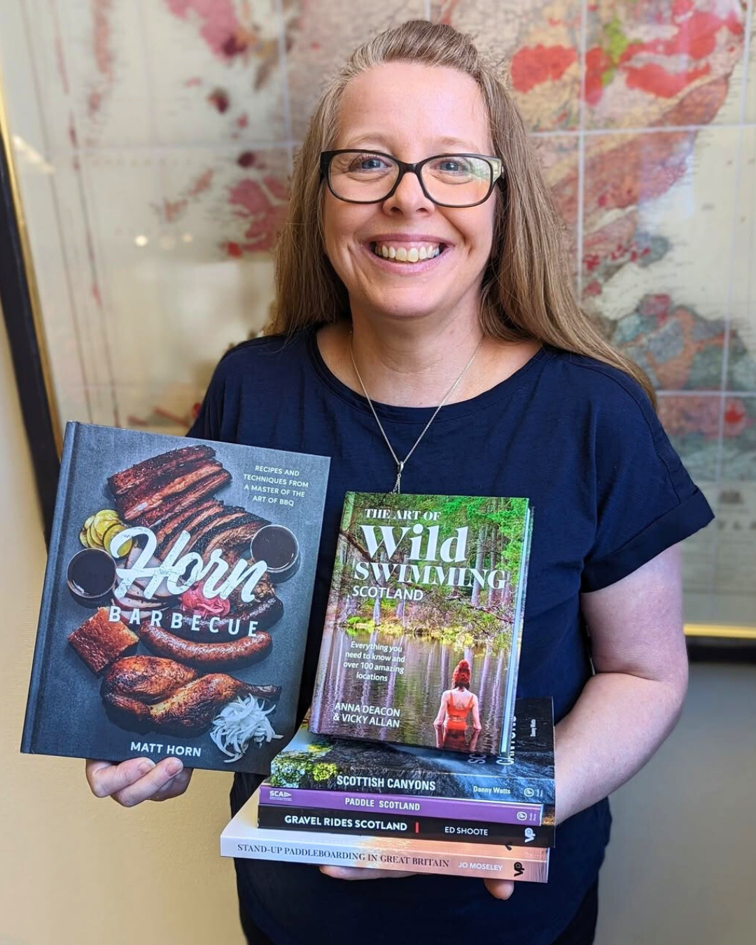 A woman with glasses smiling and holding several books about Scotland, swimming, and paddleboarding.