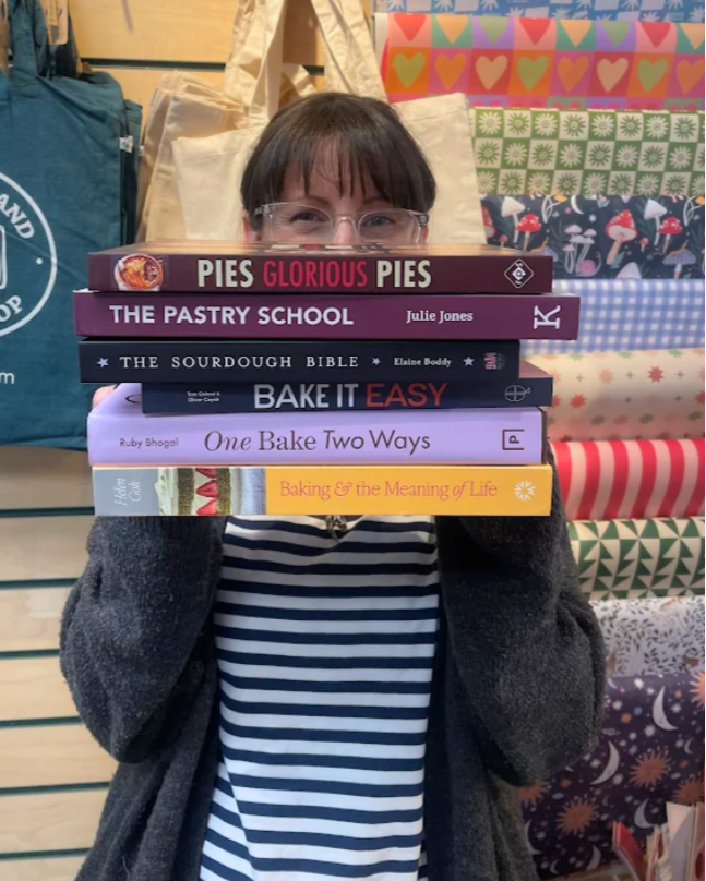 A woman with brown hair, wearing glasses, holding up a pile of cookbooks and a blue V-neck top, standing outdoors with a blurred green and brown wooded background.