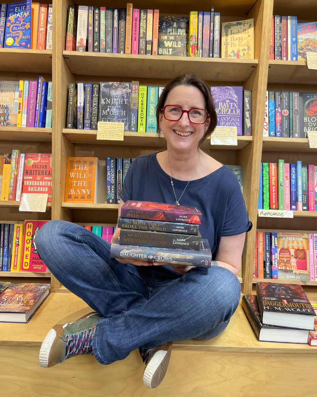 A woman with brown hair, wearing glasses, holding up a pile of books and sitting cross-legged in front of bookshelves