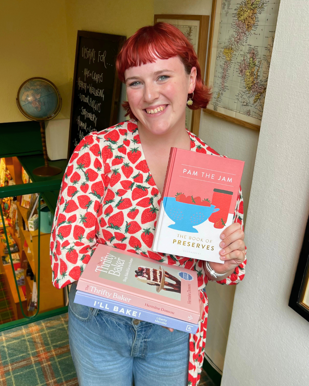 A young woman with red hair, wearing a strawberry-patterned shirt, smiling and holding two cookbooks, one titled 'Pam the Jam' and the other 'The Thrifty Baker', inside a bookstore with maps and a blackboard in the background.