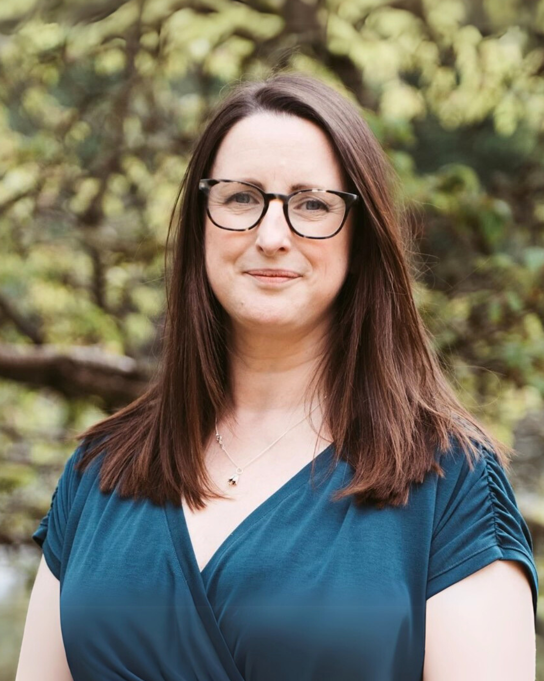 A woman with brown hair, wearing glasses and a blue V-neck top, standing outdoors with a blurred green and brown wooded background.