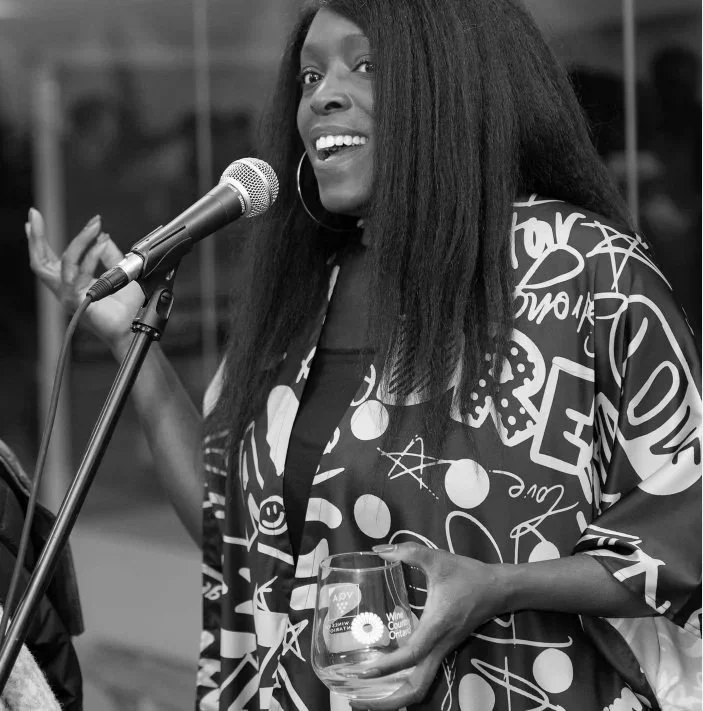 Black woman speaking into a microphone, smiling, holding a glass, wearing a patterned jacket with various doodles and symbols, in an indoor setting.
