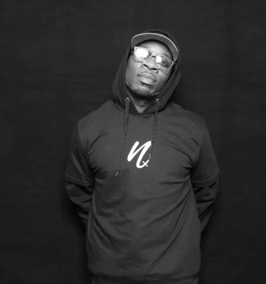 A man wearing glasses, a backwards cap, and a black T-shirt with white text poses against a black background.
