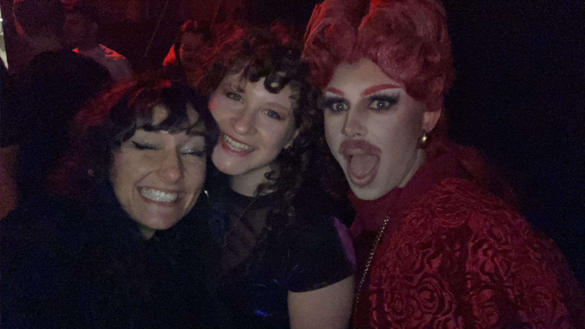 Three people smiling and posing for a photo in a dimly lit environment, one is wearing elaborate makeup and a red outfit.