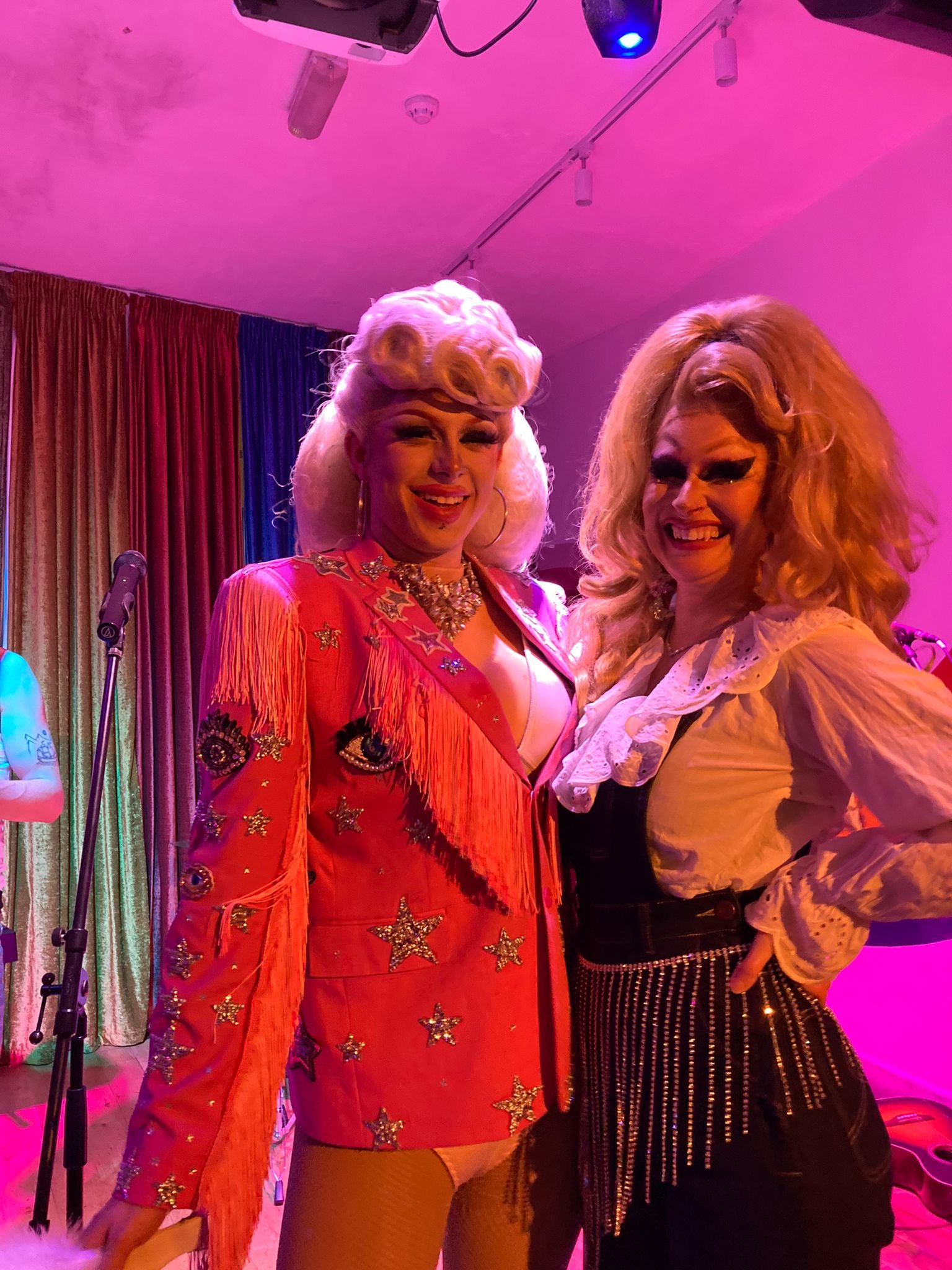Two performers in flamboyant costumes with bright makeup, standing on a stage with colorful lights.