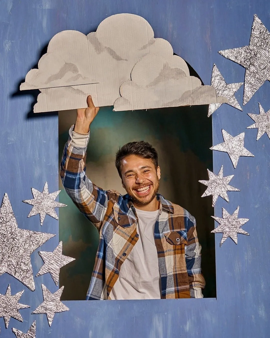 Neon Rodeo bassist Leo in a flannel shirt posing in a blue arch decorated with silver stars, holding a white cardboard cloud