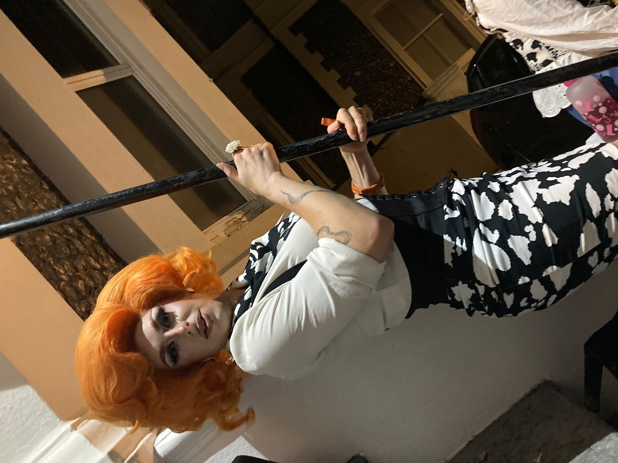 Person with orange hair and patterned outfit hanging from a horizontal pole outside, near a building wall.