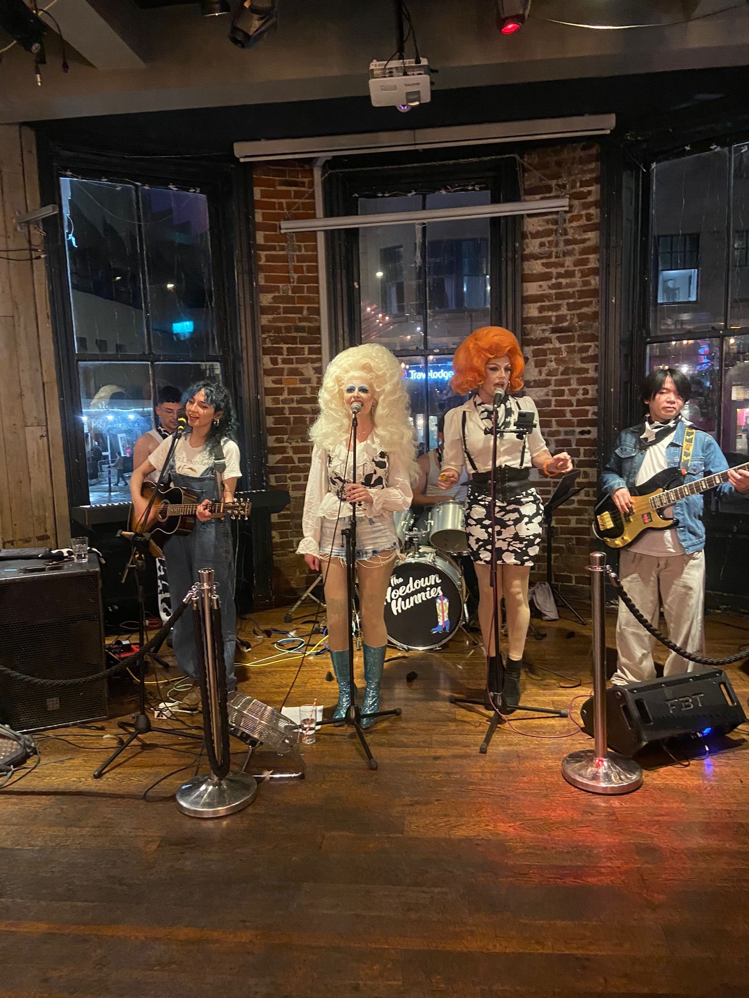 Band performing live in a venue with four musicians; two with guitars, two in colorful wigs singing into microphones, exposed brick wall background.