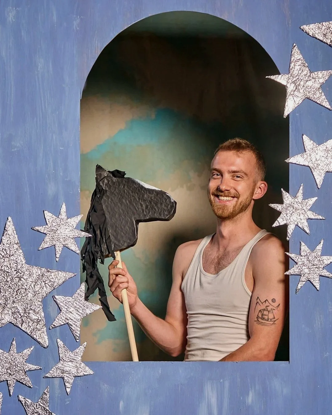 Neon Rodeo drummer Tom in a white vest posing in a blue arch decorated with silver stars, holding a hobby horse and smiling