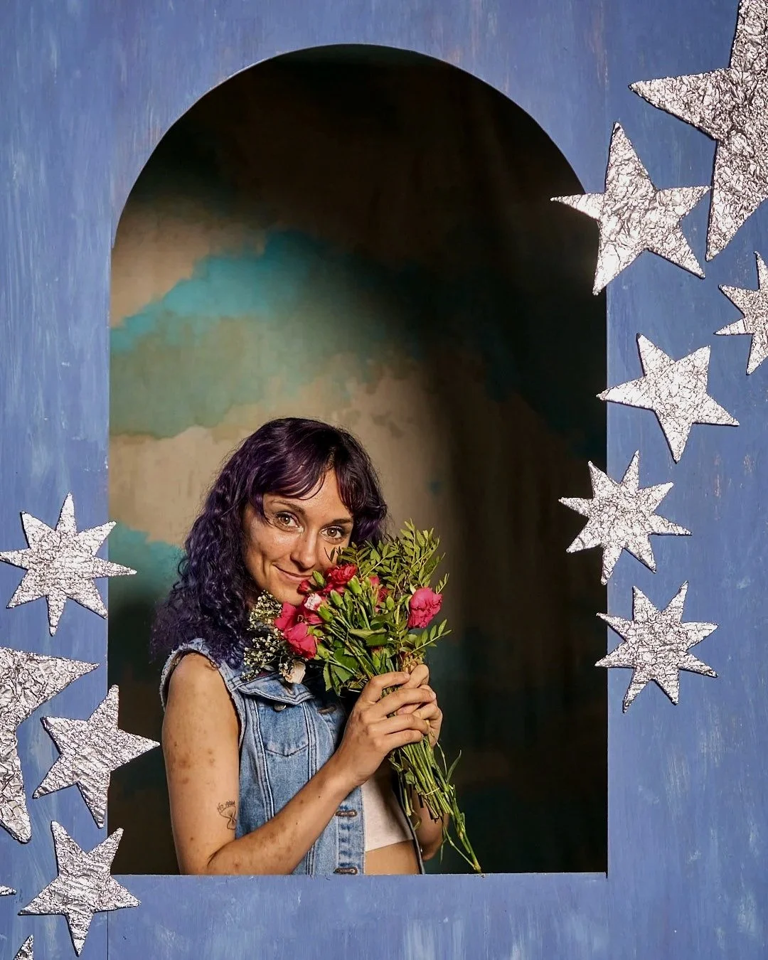 Neon Rodeo guitarist Julia Traser in a denim waistcoat posing in a blue arch decorated with silver stars, holding a bunch of pink flowers