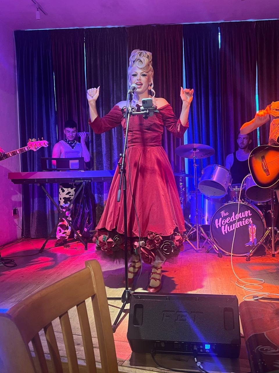 A singer in a red dress performs on stage with a band, featuring a keyboard player, guitarist, and drummer. The band name "The Hoedown Hunnies" is displayed on the drum. The stage is lit with colorful lights.