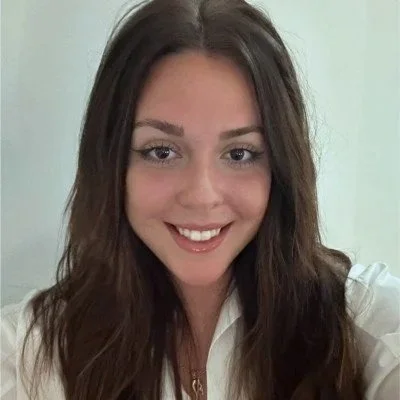A young woman with long, wavy brown hair smiling at the camera in front of a light-colored wall.