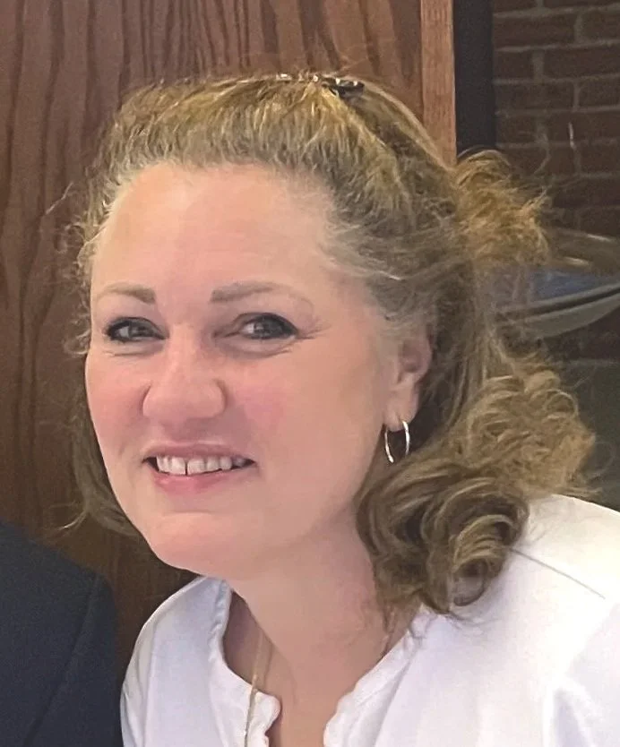 A smiling woman with curly light brown hair, wearing earrings and a white top, seated indoors with a wooden wall and brick background.