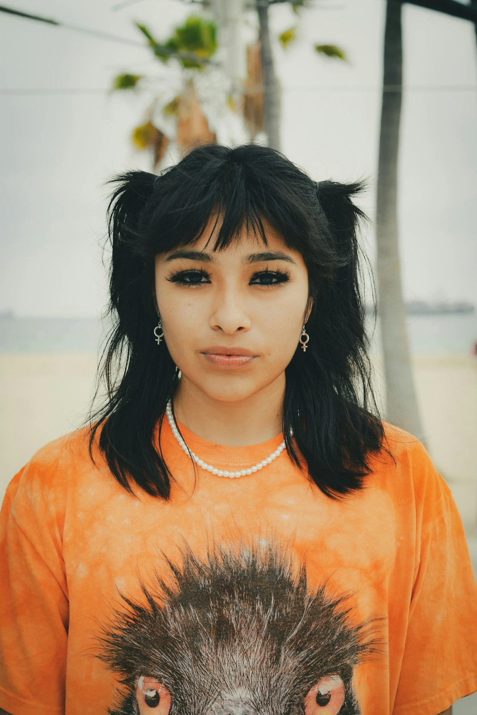 Portrait of a young woman with black hair styled in pigtails, wearing black eyeliner, a pearl necklace, earrings with female symbols, and an orange T-shirt with a lion's face on it, standing outdoors near palm trees.
