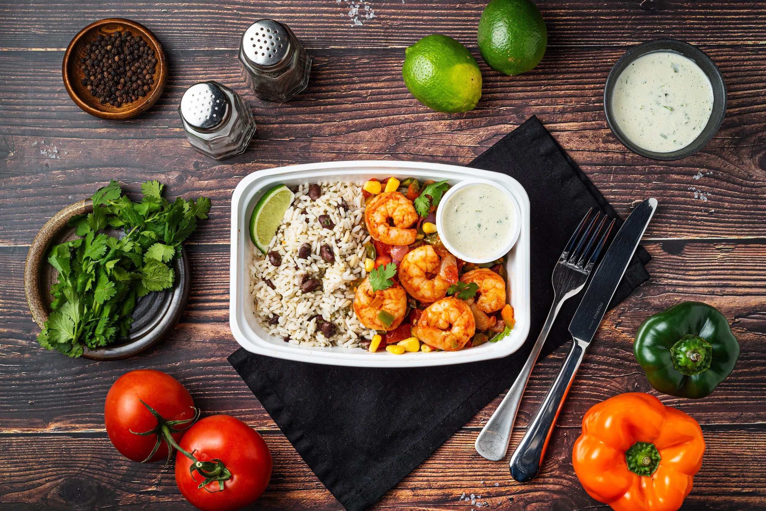 Overhead view of a meal in a white container featuring shrimp, rice with black beans, and a lime wedge. Accompanied by a small cup of sauce. Two whole tomatoes, cilantro, a green bell pepper, an orange bell pepper, peppercorns, two limes, and salt and pepper shakers are arranged around on a wooden surface. A fork and knife are placed beside a black napkin.