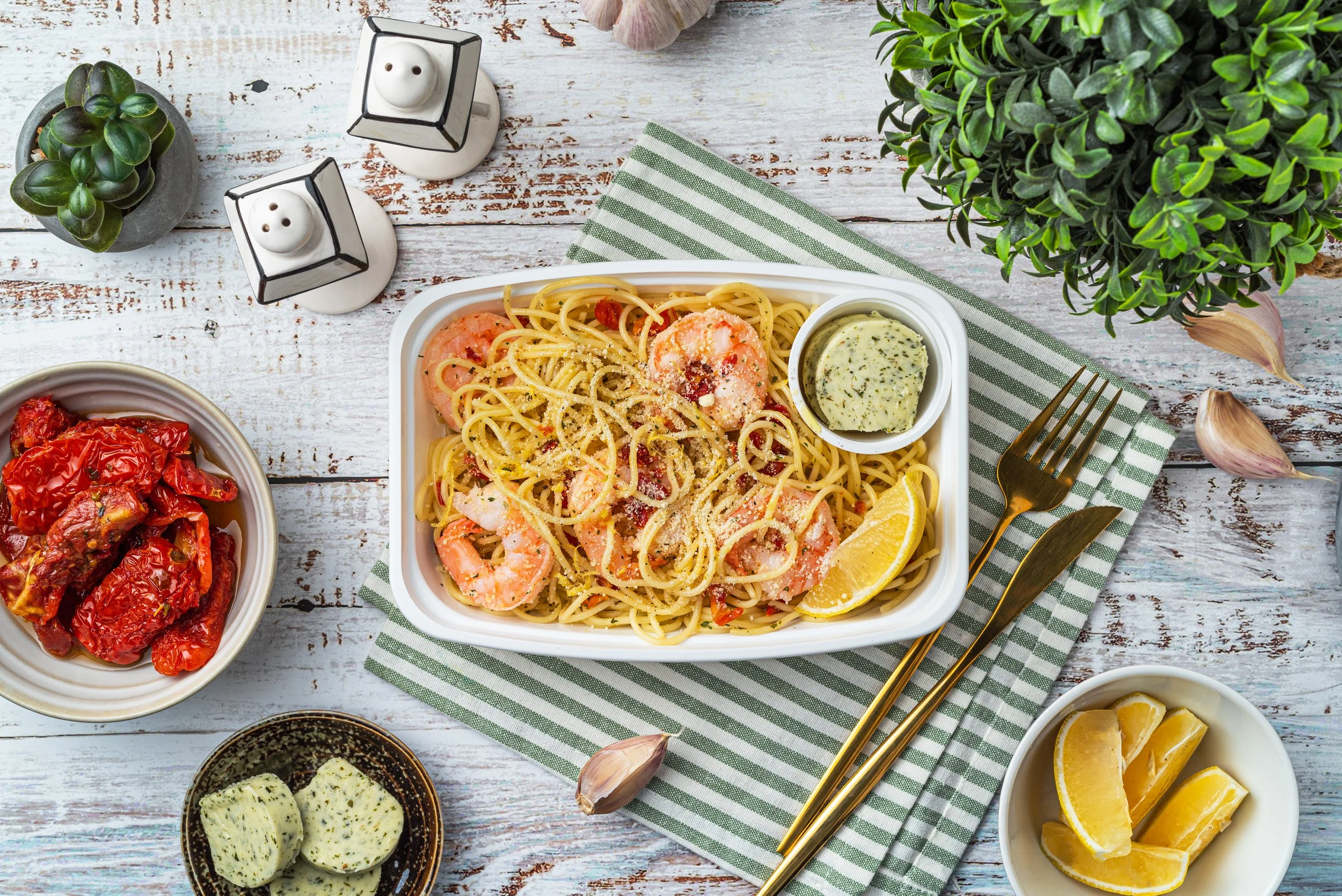 Overhead view of shrimp pasta in a white dish, garnished with lemon and herb butter, on a striped napkin. Surrounding items include a bowl of sun-dried tomatoes, lemon wedges, herb butter, salt and pepper shakers, garlic cloves, and a succulent plant.