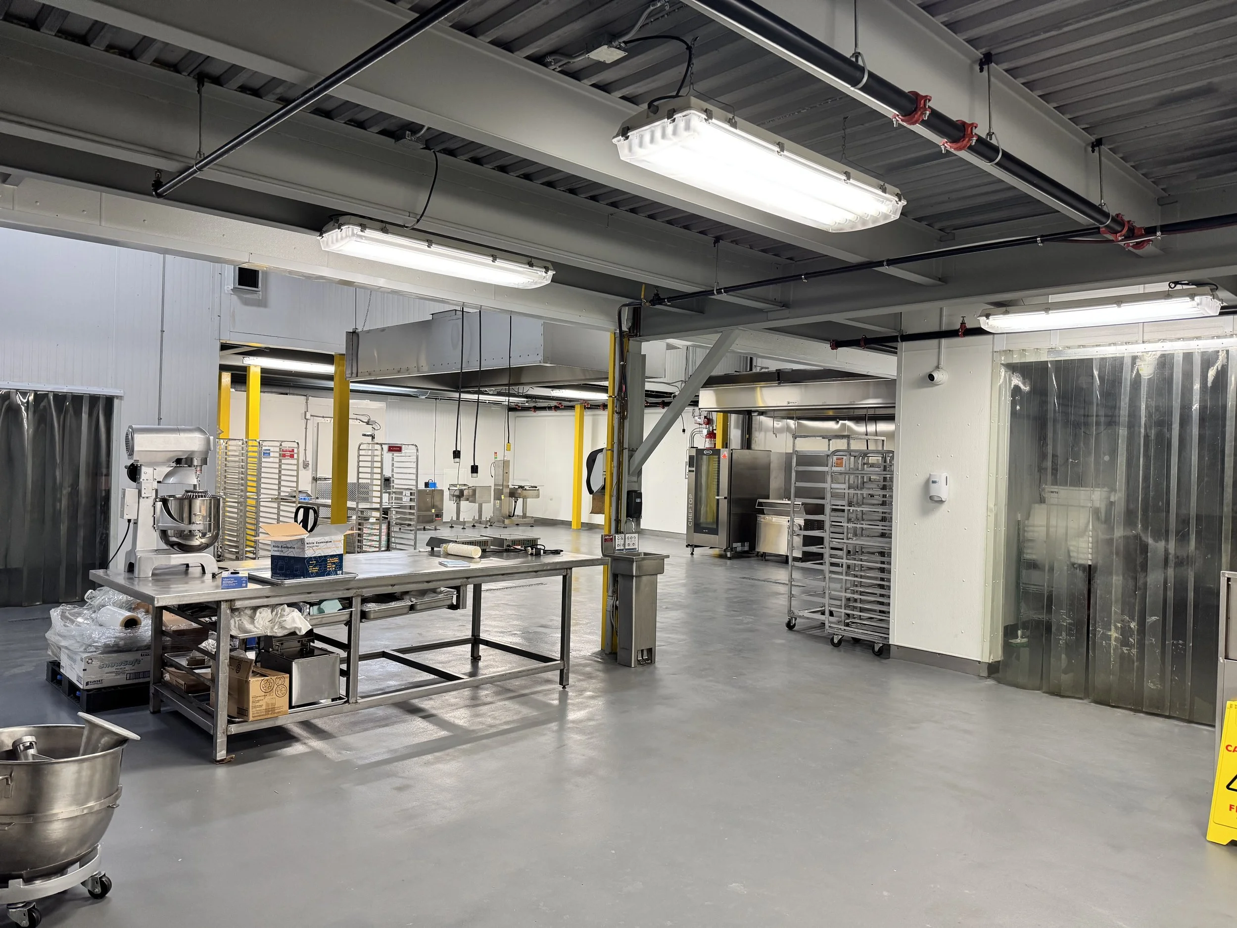Commercial kitchen with stainless steel tables, industrial mixer, rolling carts, and equipment. Overhead lighting and yellow safety posts visible.