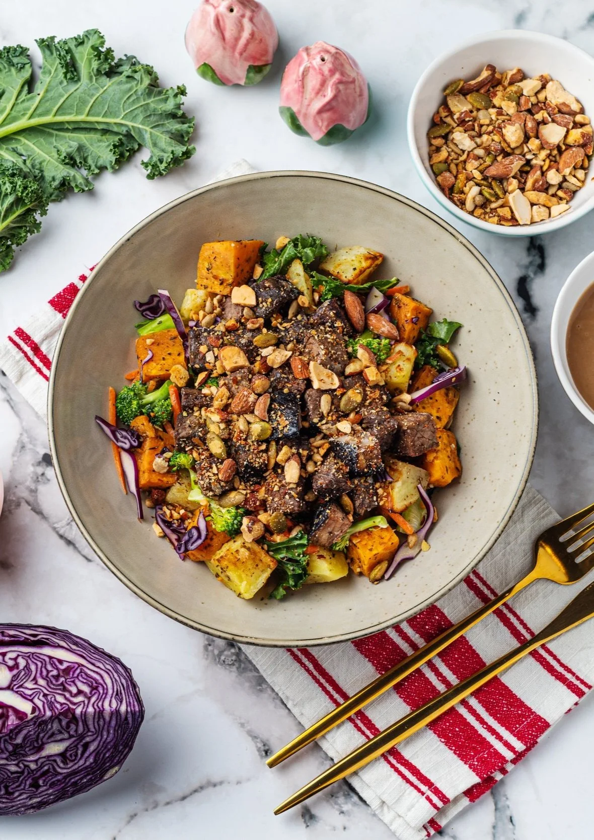 Bowl of roasted vegetables with nuts on table with kale, cabbage, utensils, and decorative shakers.