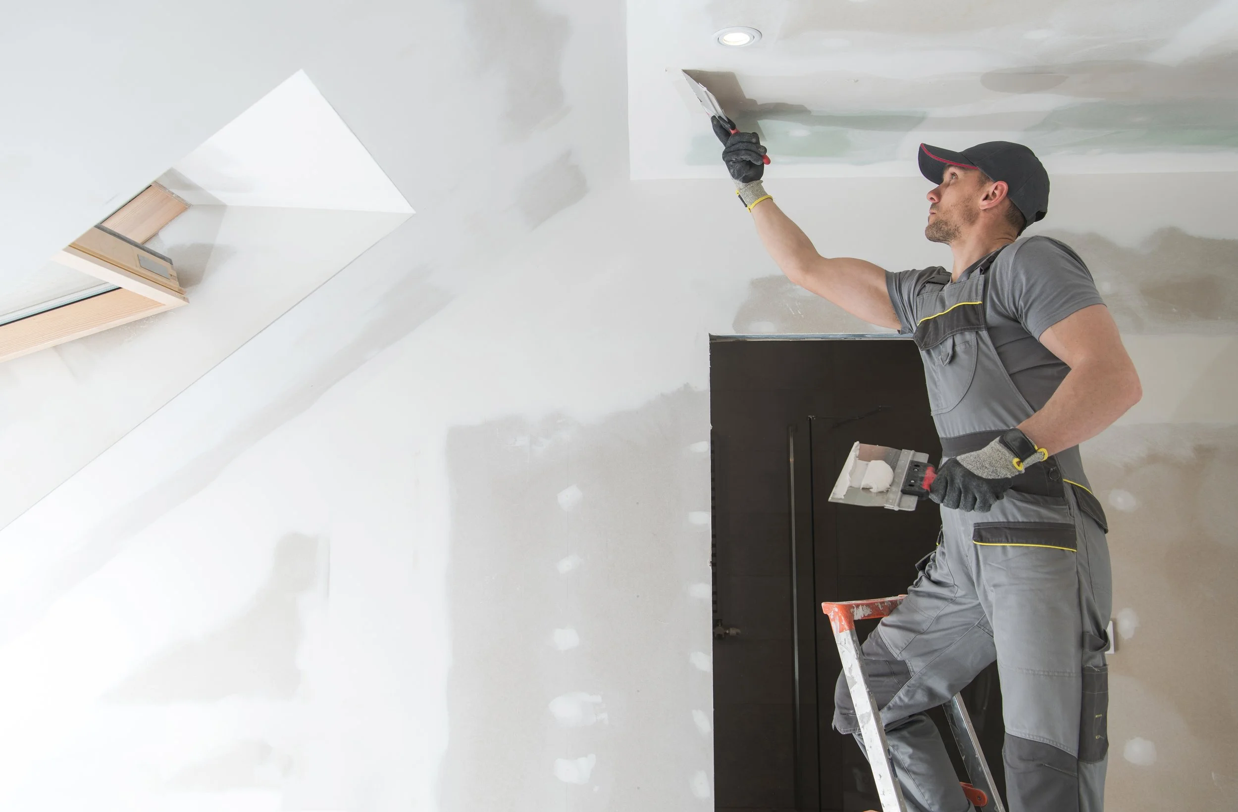 Man plastering ceiling, standing on ladder, wearing work gloves and overalls, in a room under renovation with a skylight.