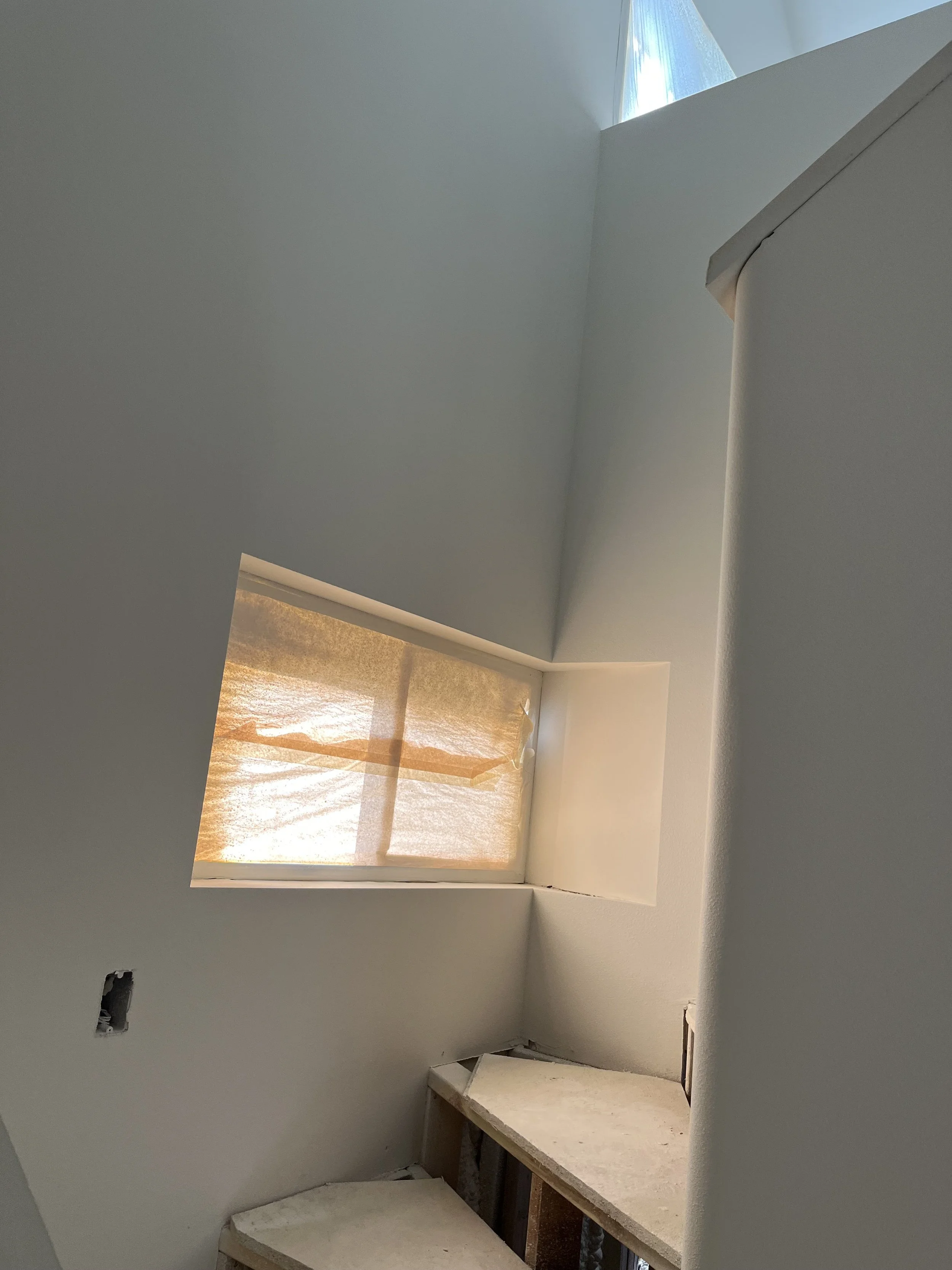 Interior of a room under construction with an angled ceiling, a small window covered with paper, and unfinished shelves underneath.
