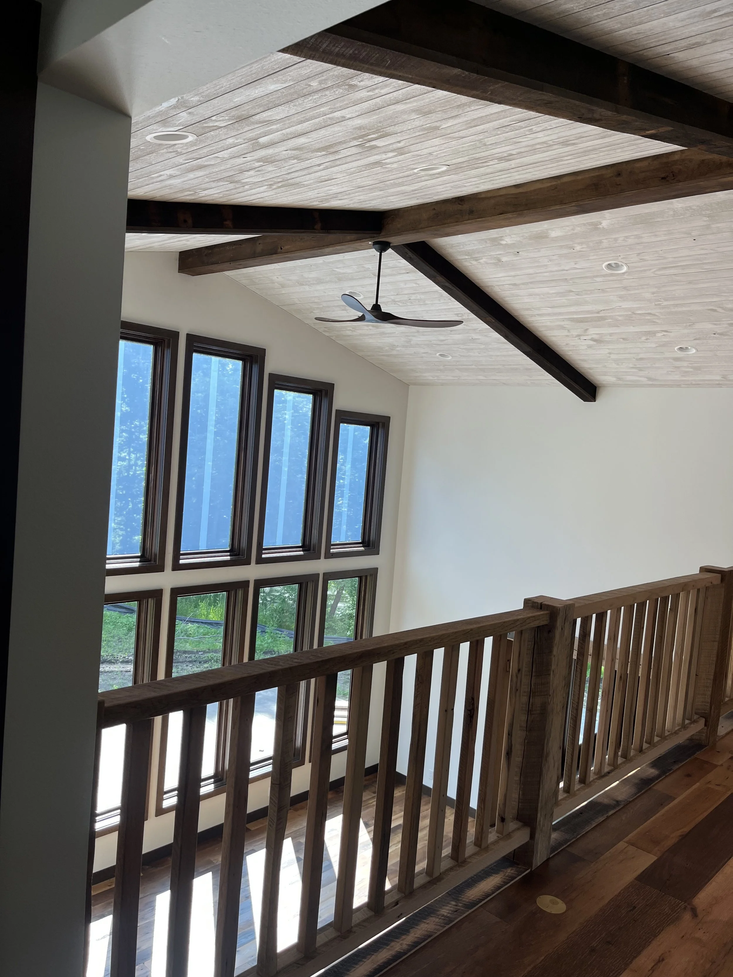 Interior view of a wooden loft with large vertical windows, a ceiling fan, and a wooden railing.