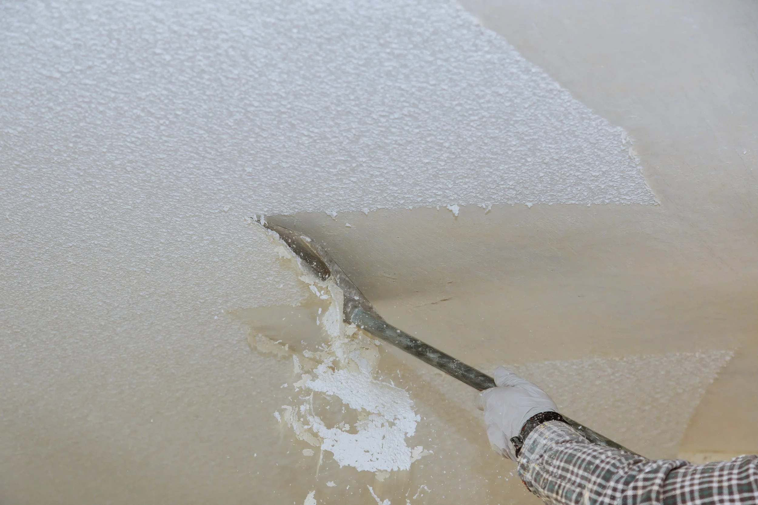 Person in a plaid shirt and white gloves removing textured ceiling with a putty knife.