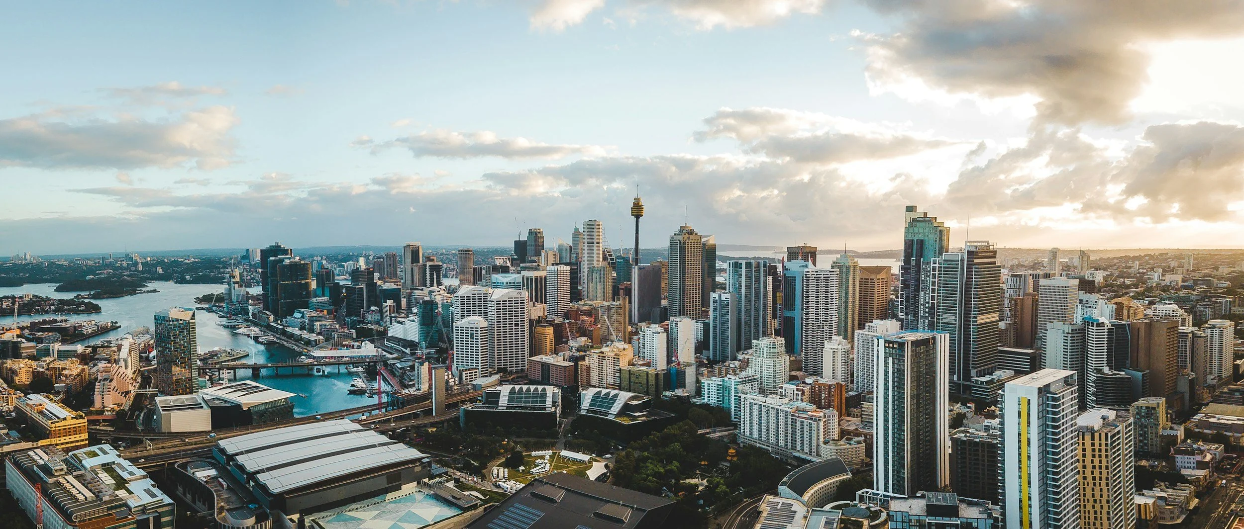 Aerial view of a modern city skyline during sunset, with tall skyscrapers, a harbor with boats, and a few clouds in the sky.