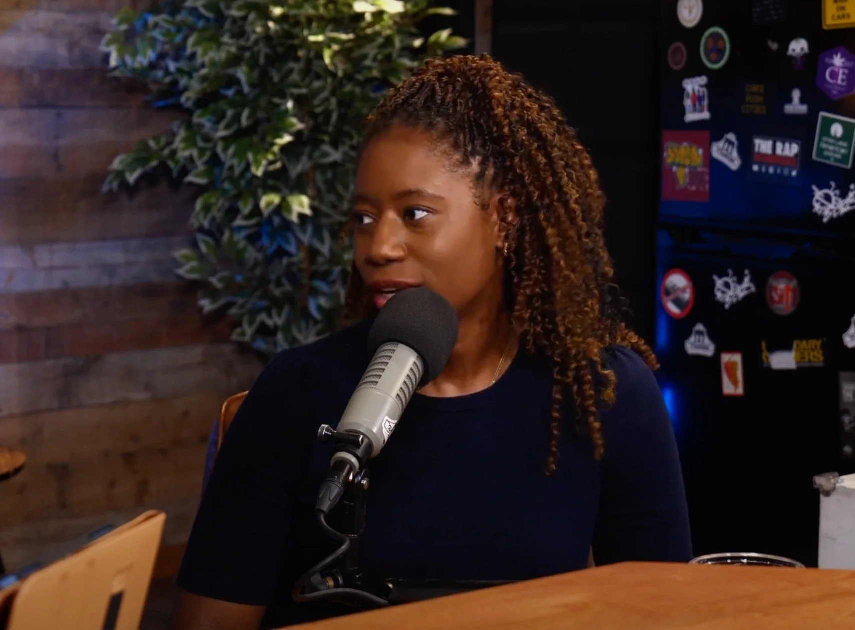 A woman with brown, curly hair speaking into a microphone in a setting with wooden wall paneling, a green leafy plant, and a black wall with various stickers.