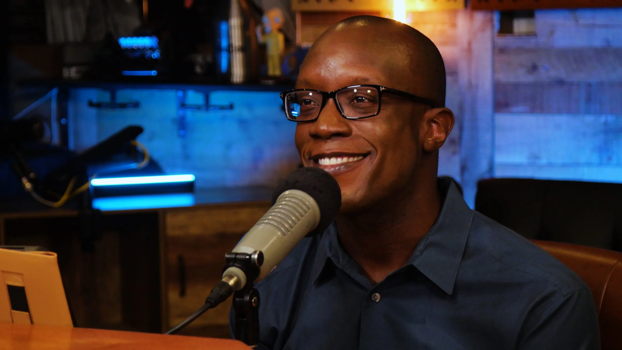 A man with glasses smiling while speaking into a microphone in a dimly lit room with a wooden background and blue neon lighting.