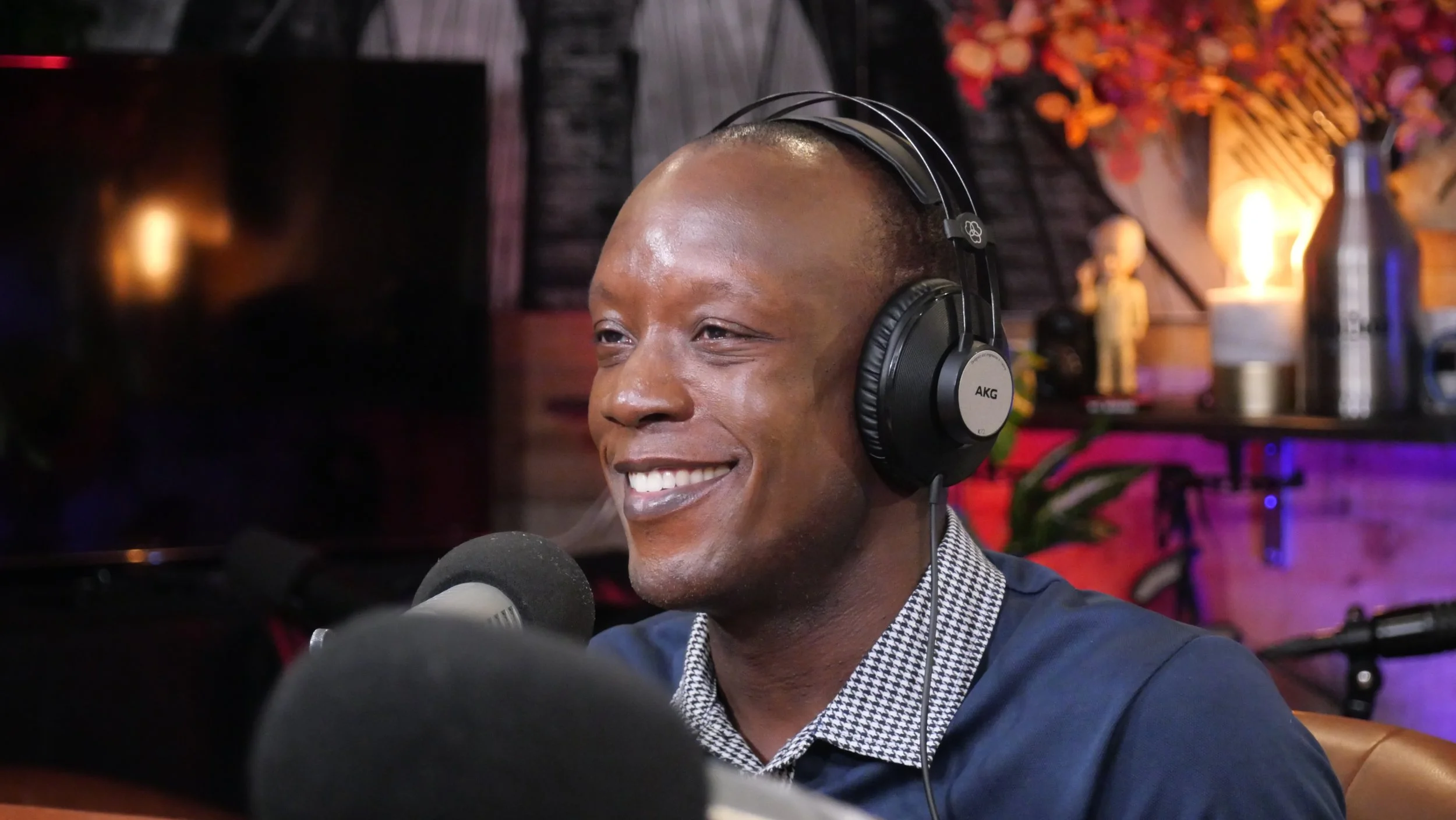 A man smiling and wearing headphones, sitting in a recording studio with dim lighting and decorative elements in the background.
