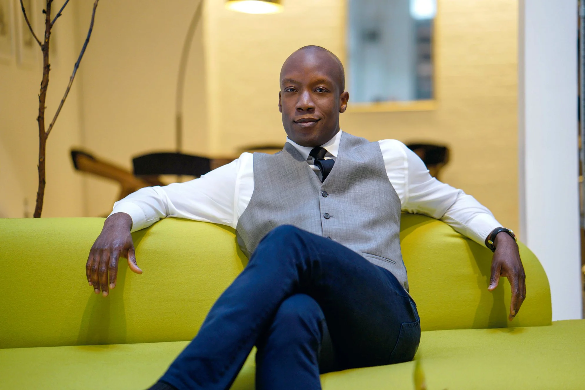 A man wearing a gray vest, white shirt, and black tie sitting on a bright yellow couch in a modern indoor setting.