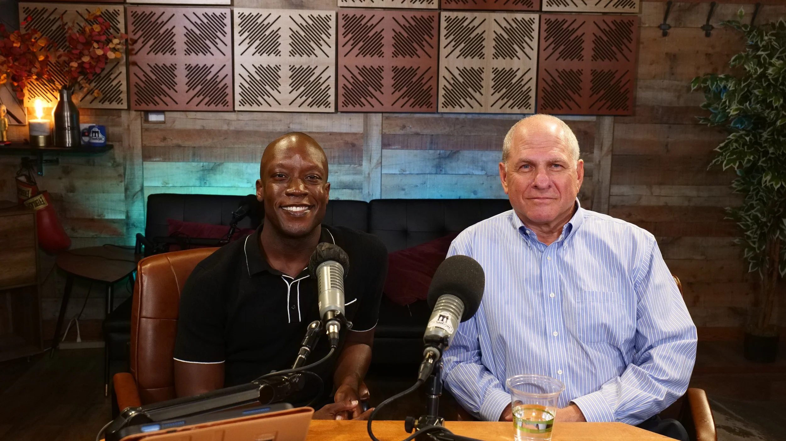 Two men sit at a wooden table with microphones, in a cozy room with wood-paneled walls, decorated with artwork and plants, appearing to be podcasting or recording.