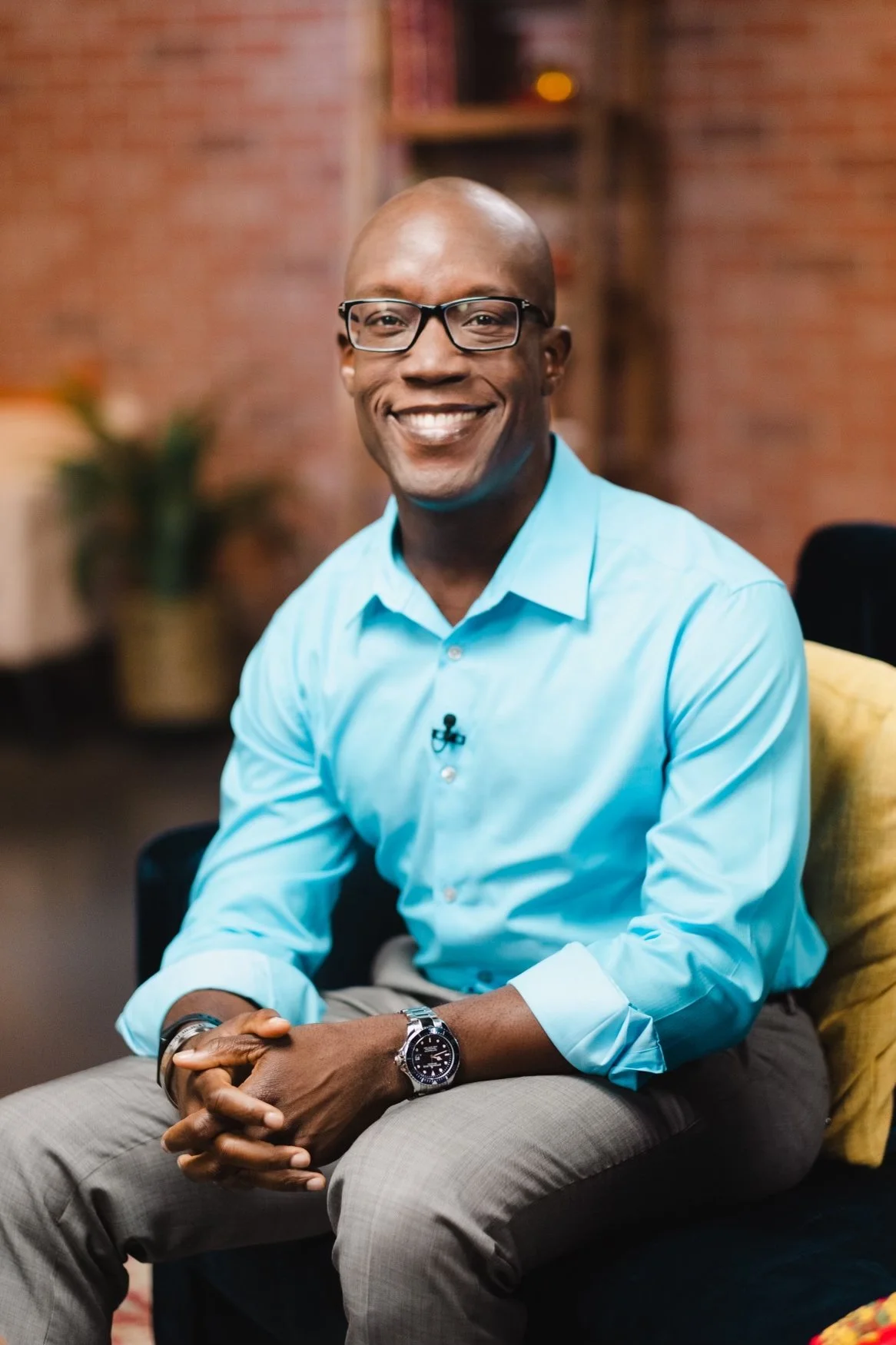 Smiling man wearing glasses, a blue dress shirt, and a watch, sitting in a studio with a brick wall background and a bookshelf.